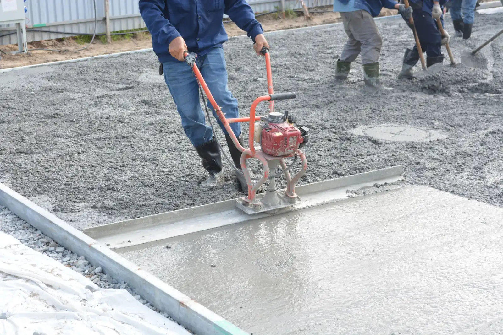 Worker running a power screed across a freshly placed concrete slab while others rake behind him