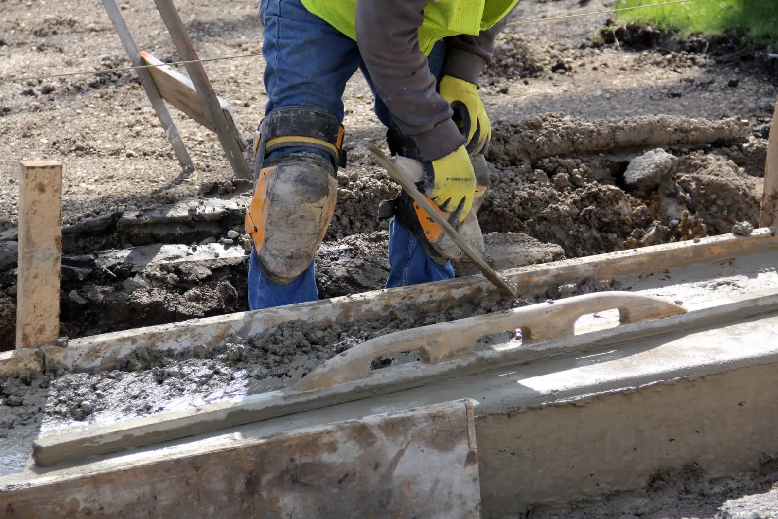Worker in knee pads hand-tooling the edge of a fresh concrete slab along a wooden form board