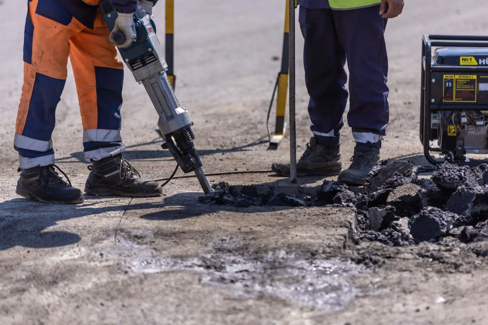 Two workers in high-visibility clothing breaking up concrete pavement with a jackhammer