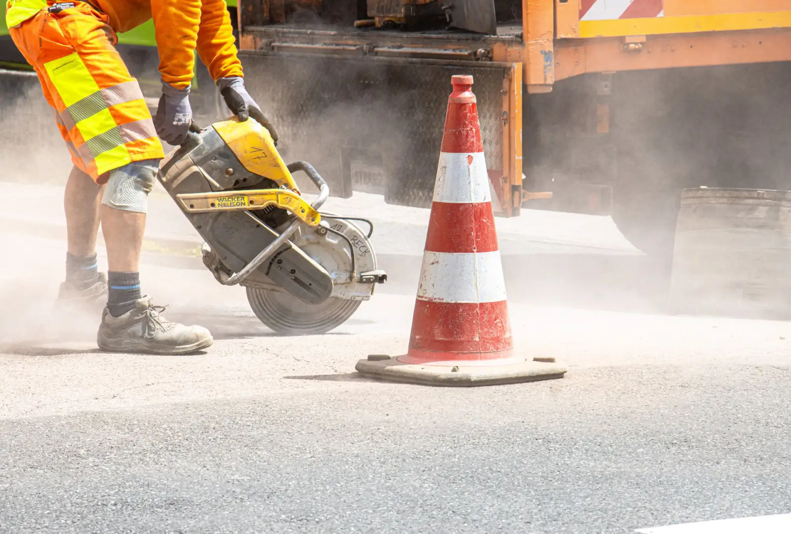 Worker running a walk-behind concrete saw to cut pavement beside a traffic cone
